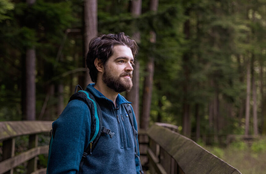 A bearded man standing on a wooden bridge in the woods.
