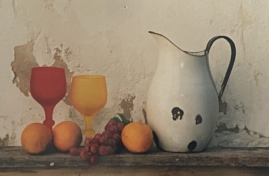 A pitcher with oranges and grapes on a table.