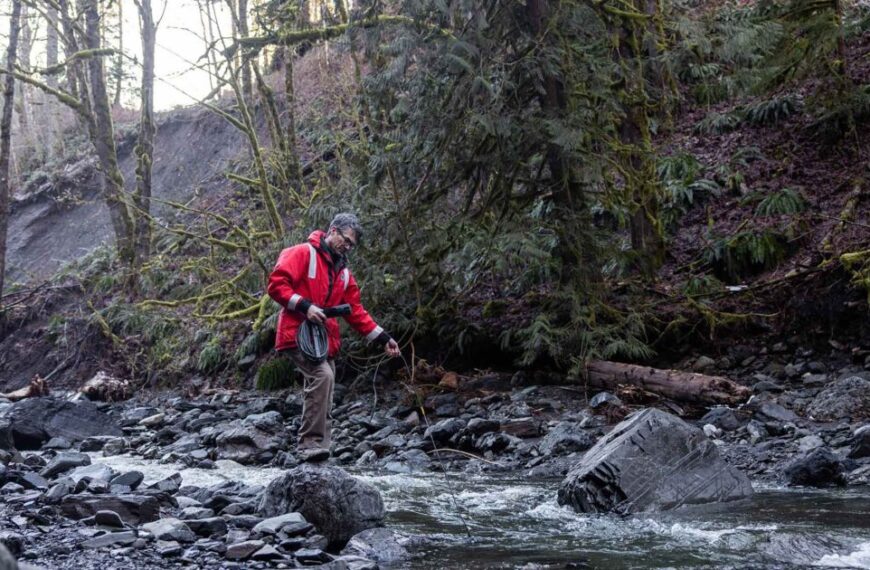 A man in a red jacket is crossing a stream.