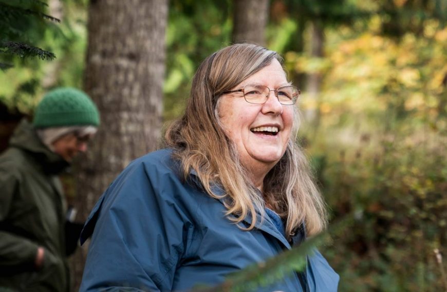 A woman in a blue jacket is smiling in the woods.