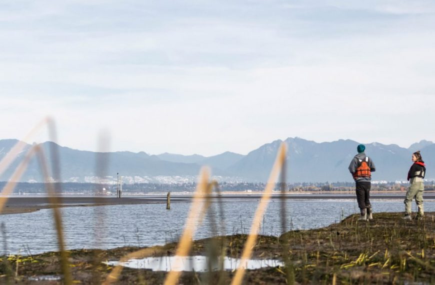 Two people standing on the shore of a body of water with mountains in the background.