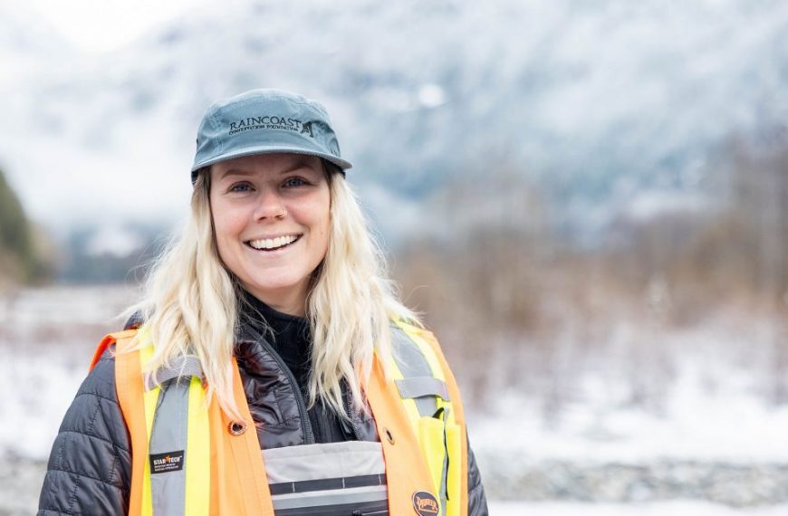 A woman in a safety vest smiling in front of mountains.