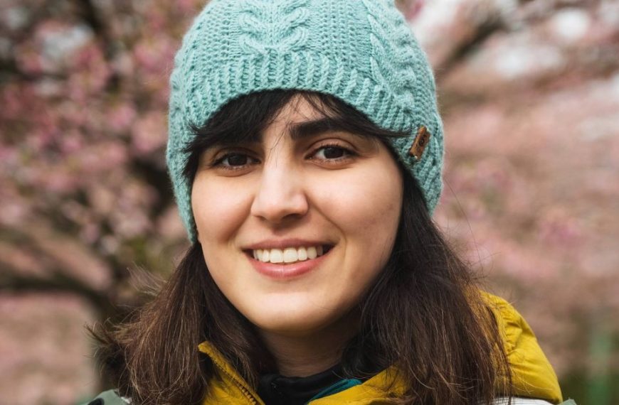 A young woman wearing a blue beanie in front of a blossoming cherry tree.