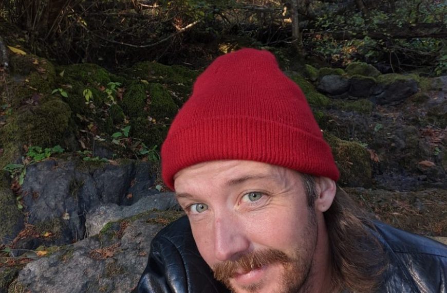 A man in a red beanie taking a selfie in the woods.