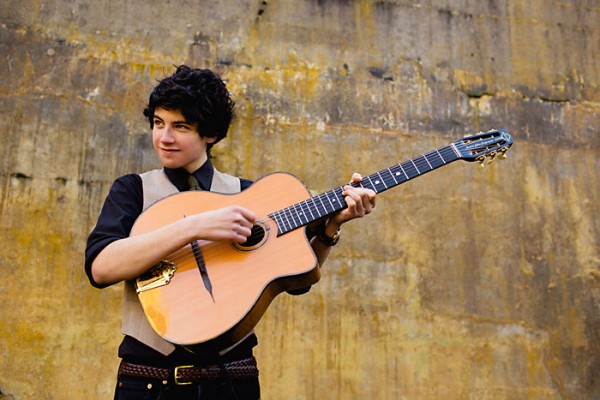 A young man holding an acoustic guitar in front of a wall.