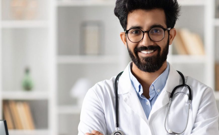 A male doctor in a lab coat standing in front of a computer.