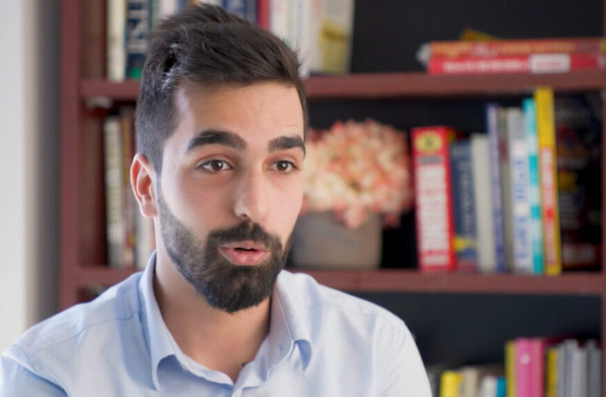 An employed Syrian man with a beard standing in front of a bookcase, highlighting the integration of refugees into the workforce.