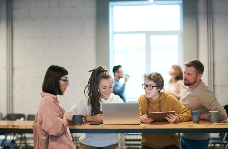 A group of people sitting around a table, discussing and glancing at a laptop screen.