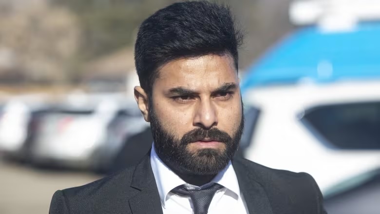 A man, possibly Jaskirat Singh Sidhu, dressed in a suit and tie is seen standing in a parking lot.