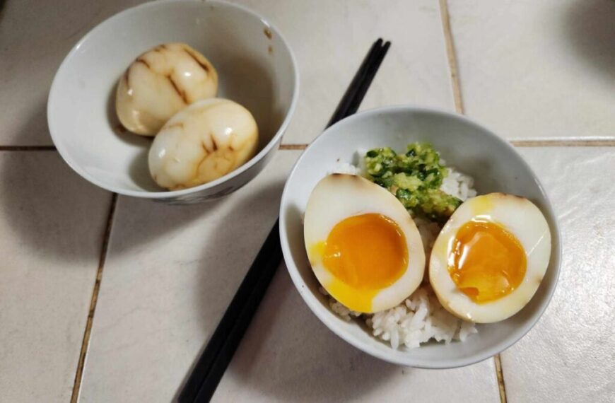 Two bowls containing food items on a tiled surface with a pair of black chopsticks resting beside them. The left bowl has two H&H Chinese Tea Eggs, and the right bowl contains rice topped with