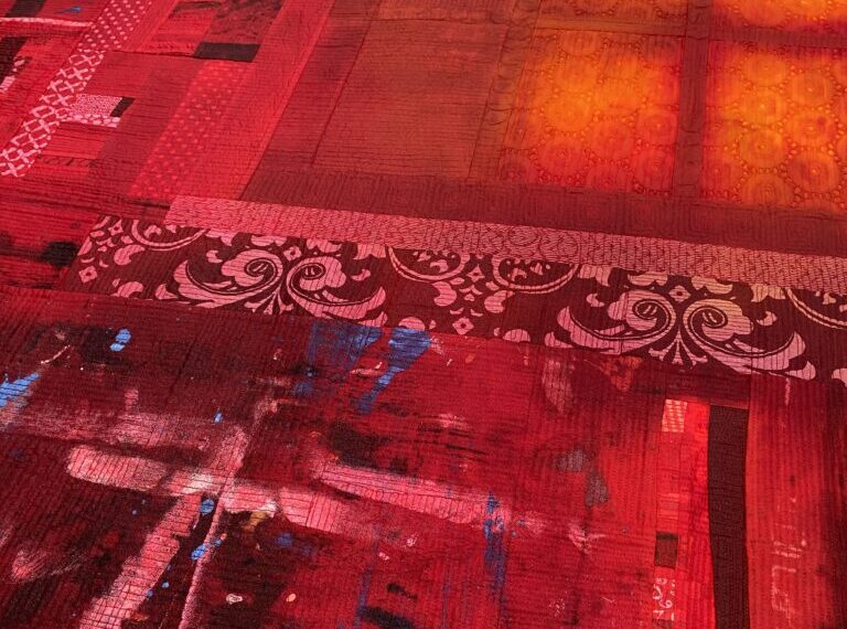 A close-up of a colorful quilt against a bare design wall, prominently featuring red patches with various intricate patterns and textures.
