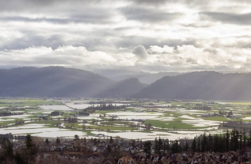 Aerial view of a scenic valley with fields partially flooded, surrounded by hills under a cloudy sky with sun rays breaking through. A small plane is visible in the upper left corner, symbolizing the ongoing efforts toward ecosystem restoration and reconciliation at Sumas Xhotsa.