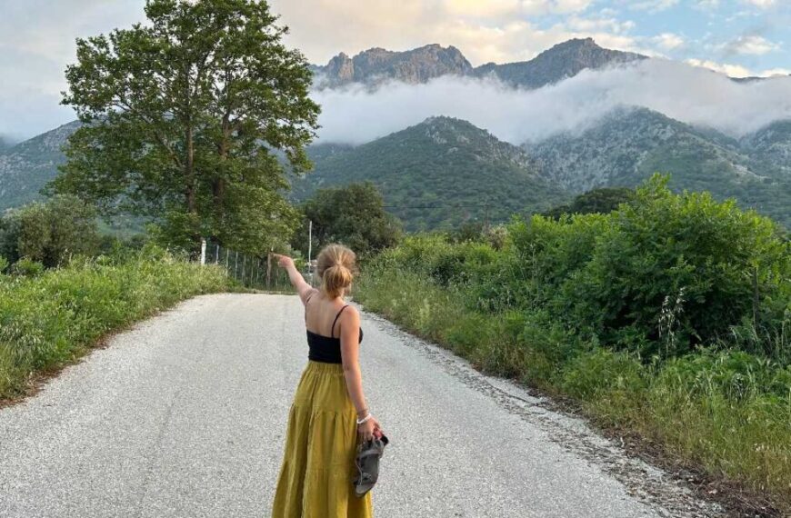 Olivia Tomlinson, wearing a yellow skirt and black top, stands on a gravel road pointing toward distant mountains partially covered by clouds, surrounded by lush greenery.