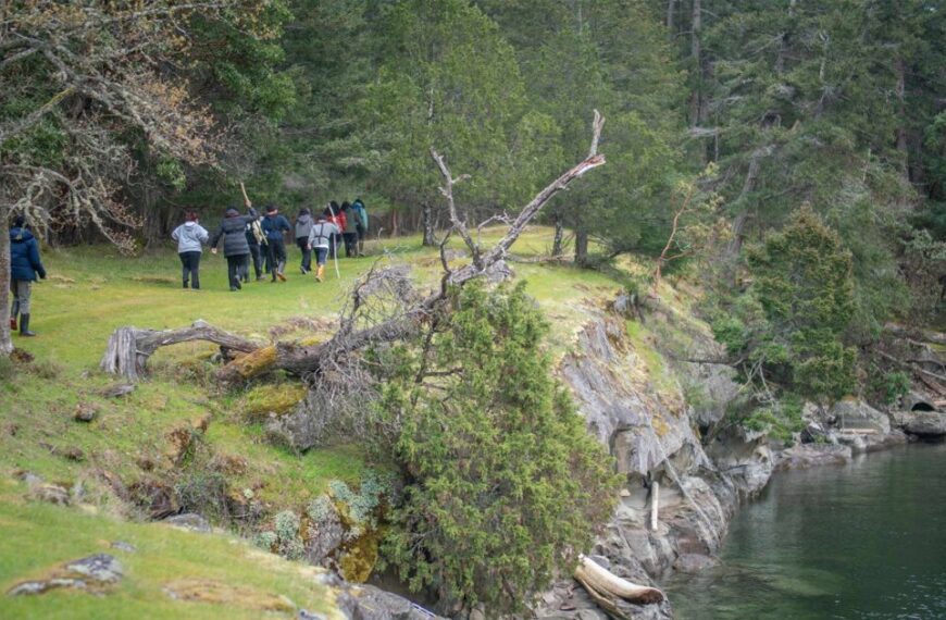 A group of people, known as Emerging Stewards in their eighth year, walk along a grassy path beside a rocky cliff edge with trees and the serene Salish Sea in the background.