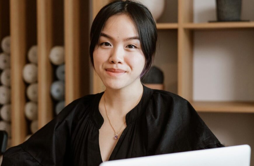 A woman with black hair, wearing a black top, sits in front of a laptop, smiling slightly as she reviews the latest updates on the Interconnectedness Initiative. Shelves with neatly arranged items are visible in the background.