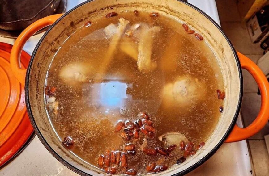 A pot with chicken drumsticks, red beans, and other ingredients simmering in an Herbal Broth on a stove. The pot has orange handles and lid, perfect for Fall Immune Support.