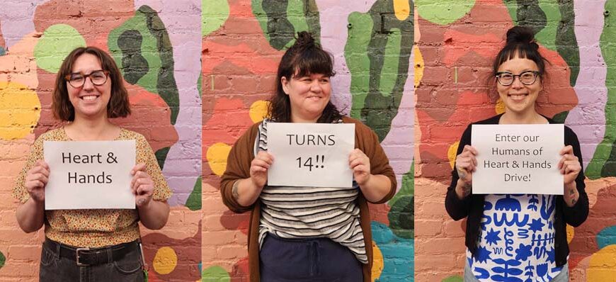 Three individuals stand against a colorful brick wall holding signs that read: "Heart & Hands," "H&H turns 14!!," and "Enter our Humans of Heart & Hands Drive!" respectively.