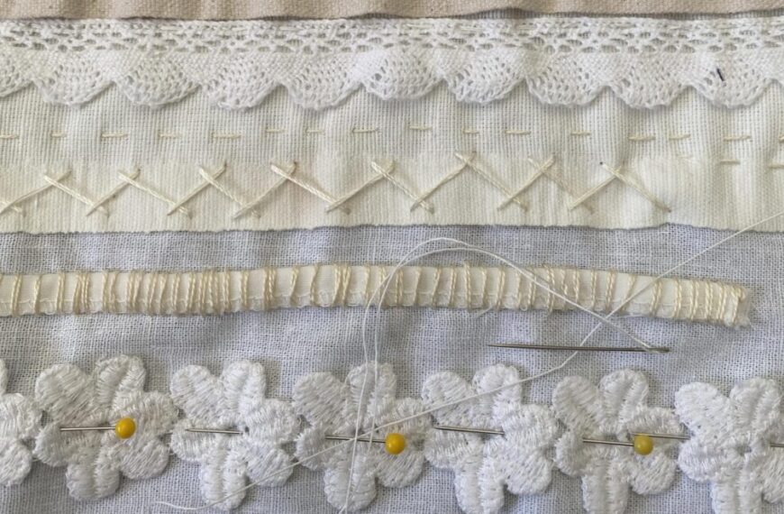Close-up of white fabric with various patterns of hand-stitched designs, including lace, crisscross stitches, a gathered stripe, and flower shapes pinned with yellow-headed needles and threaded scraps.
