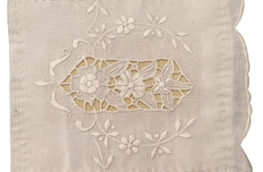 A rectangular piece of white fabric features intricate floral embroidery and cutwork designs with scalloped edges on the right side, showcasing the art of detailed stitching.