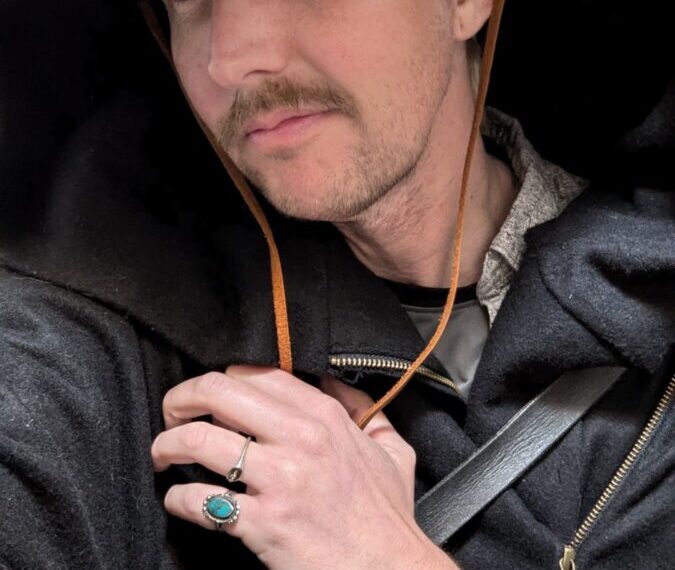 A person wearing a wide-brimmed hat, black cloak, and a ring with a green stone looks at the camera, seemingly ready for updated events. Their left hand is raised to their chest.