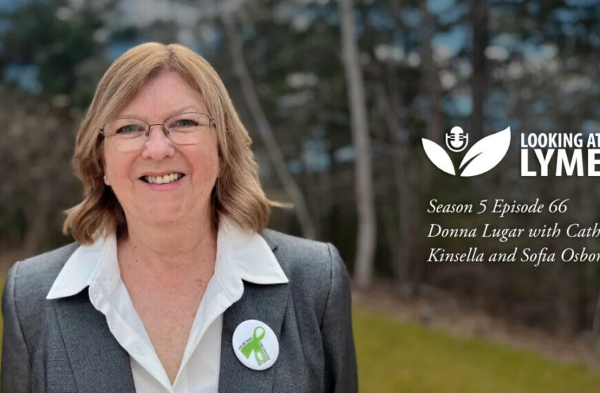 A person smiling outdoors, wearing glasses and a gray blazer adorned with a Lyme disease awareness pin. With "Looking at Lyme" Season 5, Episode 66 featuring Donna Lugar, this episode focuses on advocating for change in Lyme disease advocacy.