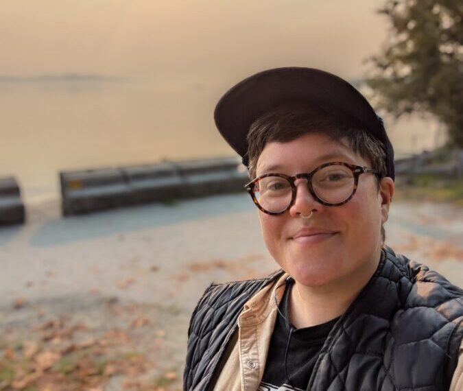 A person with glasses and a cap smiles at the camera near a hazy waterfront, promoting inclusion as the sun dips low in the sky.