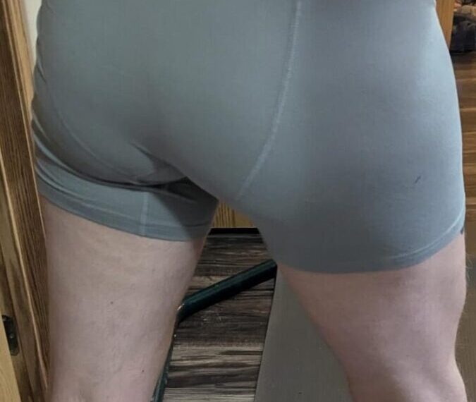 A person in gray shorts stands with their back to the camera, partially visible in a reflective surface, possibly contemplating a game of boule.
