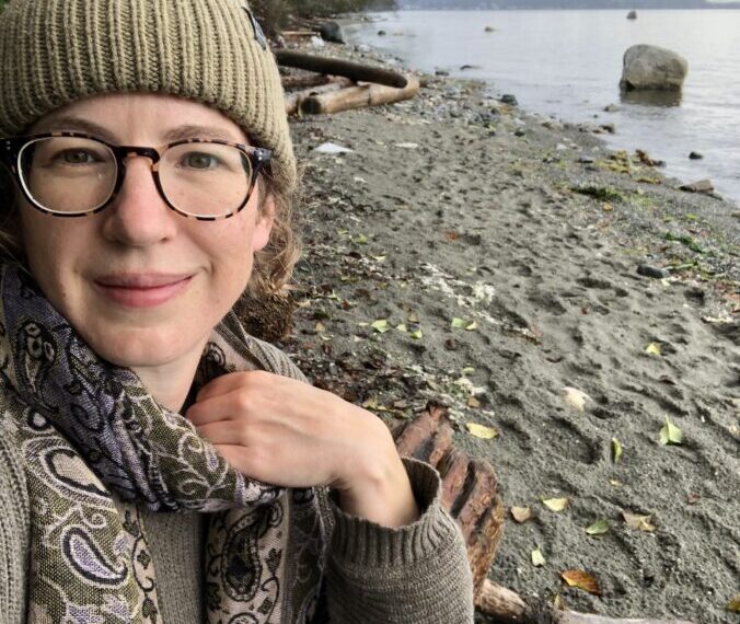 A person wearing glasses, a beanie, and a scarf sits on a rocky beach with driftwood and calm water in the background, embodying the quiet wisdom of nature's serene beauty.
