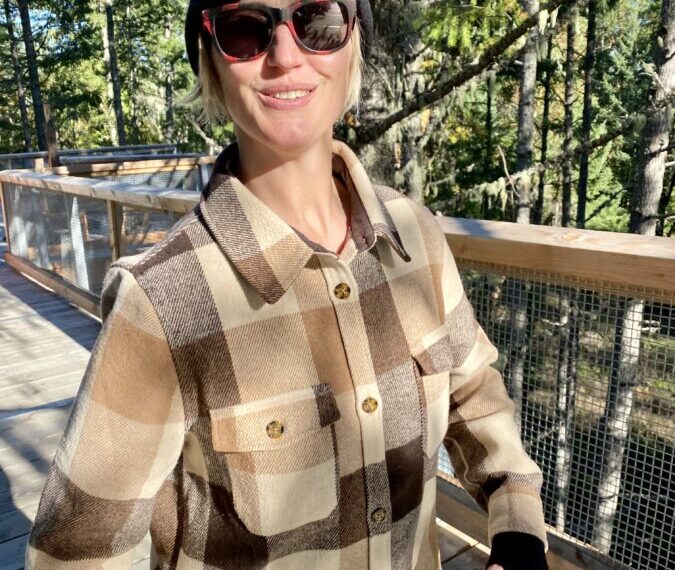 A person wearing a plaid jacket, beanie, and sunglasses stands on a wooden walkway in a forested area, embracing the tranquility of nature as part of their self-care routine from Heart & Hands.