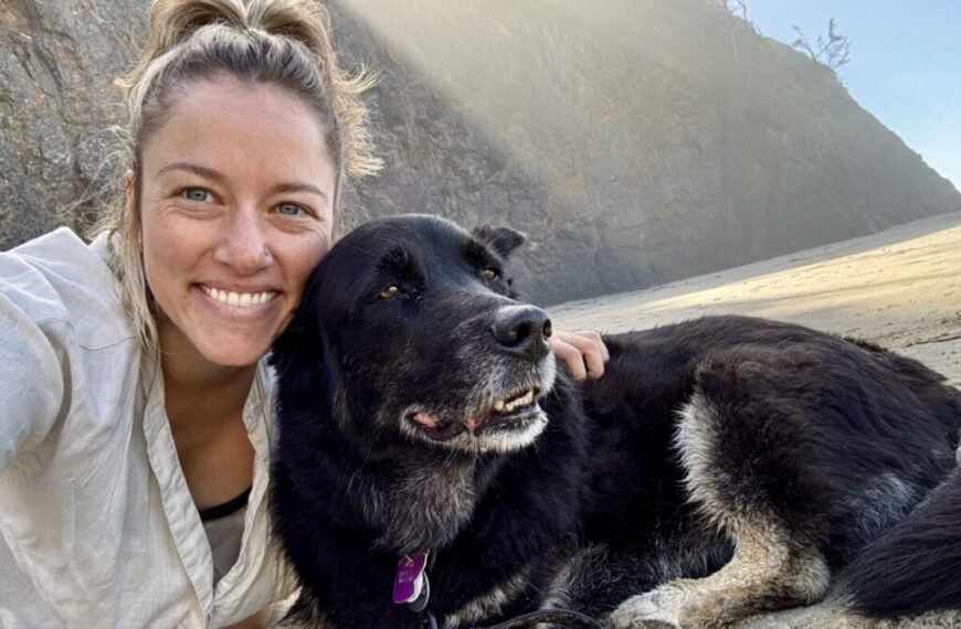 A woman and a black dog sit on a sandy beach with rocky cliffs in the background, both looking content, embodying the peace found at Heart & Hands—a clinic known for its accessible care.