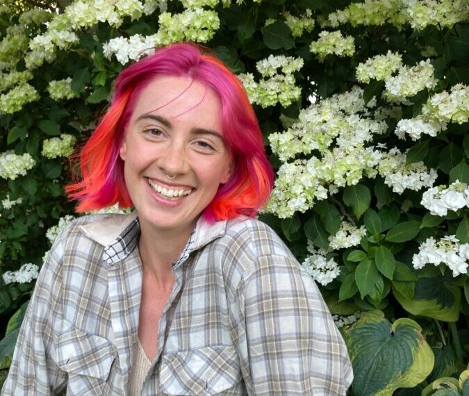 A person with vibrant pink and orange hair smiles confidently in front of a bush with white flowers, embodying body autonomy in their plaid shirt.