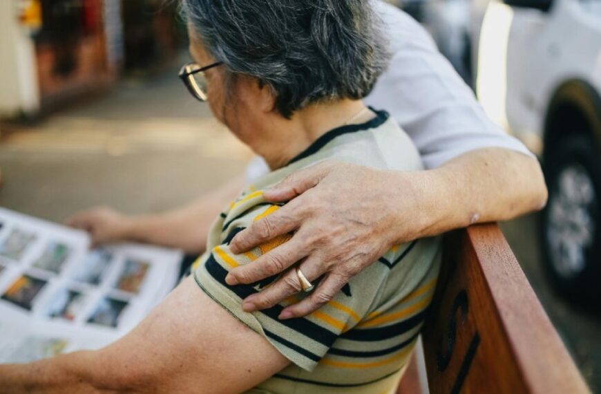 Two elderly individuals sit on a bench; one reads a book, while the other places a hand on their shoulder. A parked white vehicle is visible in the background. This heartfelt moment reflects the spirit of October 1, International Day of Older Persons.