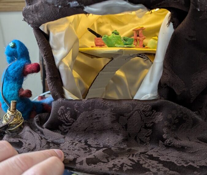 A small diorama scene with a window display reveals miniature figures amidst props, evoking musings of tiny worlds. A hand slowly lifts the fabric cover, unveiling this STICC-inspired creation ready for imaginative role play.