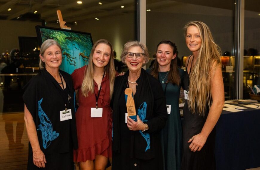 Five women stand together, smiling at an evening indoor event, with art displayed in the background. One woman proudly holds a 2024 Raincoast Ocean Science Award.