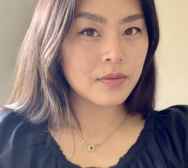 A person with shoulder-length dark hair wearing a black top and a necklace with a life-affirming green pendant looks into the camera.