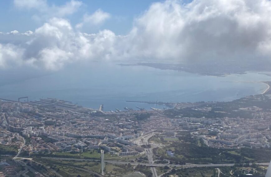 Aerial view of a coastal city in Portugal, with visible docks, shimmering waters, buildings, and lush green spaces. Clouds are scattered above the scene under a blue sky, capturing the soul of Portugal with its breathtaking landscape.
