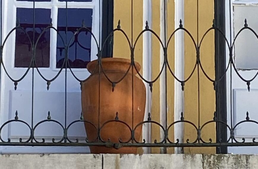A terracotta pot graces a balcony with black wrought iron railing, echoing the timeless traditions of Évora against a yellow and white building facade with windows.