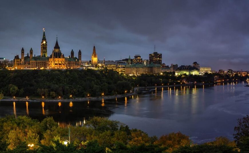 Night view of Ottawa's skyline with Parliament Hill, the heart of the federal government, illuminated and reflecting on the Ottawa River under a cloudy sky.