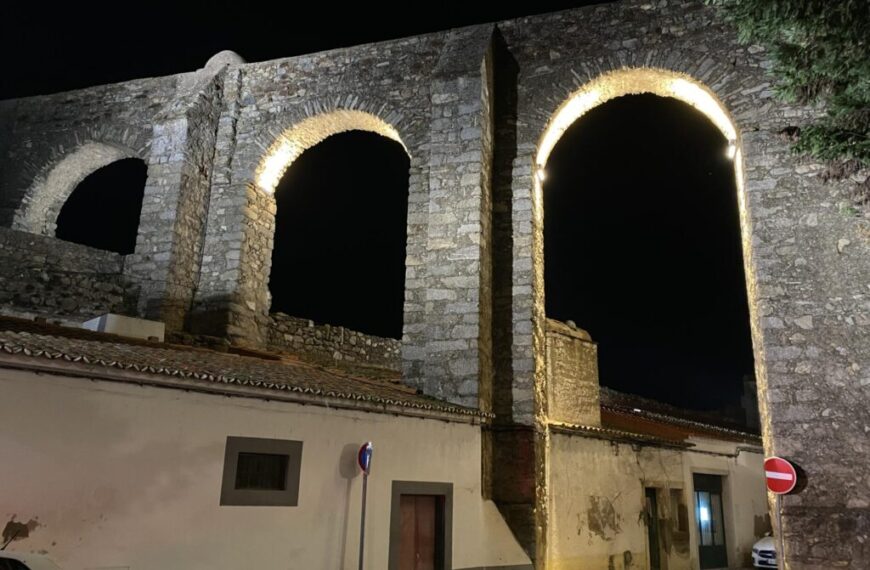 Stone aqueduct arches lit at night, casting a warm glow over the scene; a building stands below, welcoming travelers and homebodies alike. Signs guide the wandering spirit of a travelling soul while embracing the comfort of a homebody's heart.