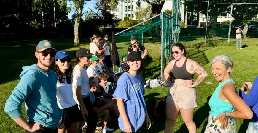 A group of people, some standing and others sitting on the grass near a green fence in Oak Bay, are smiling in the sunny park, enjoying their time together.