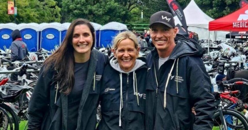 Three people, possibly clients or physios, smiling in front of a park event with bicycles and tents in the background, wearing jackets.