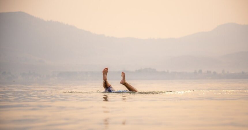 A person floats on their back in the calm waters of Elk Lake, legs gracefully raised above the surface. Hazy mountains provide a serene backdrop, reminiscent of a peaceful moment during the Elk Lake Triathlon.