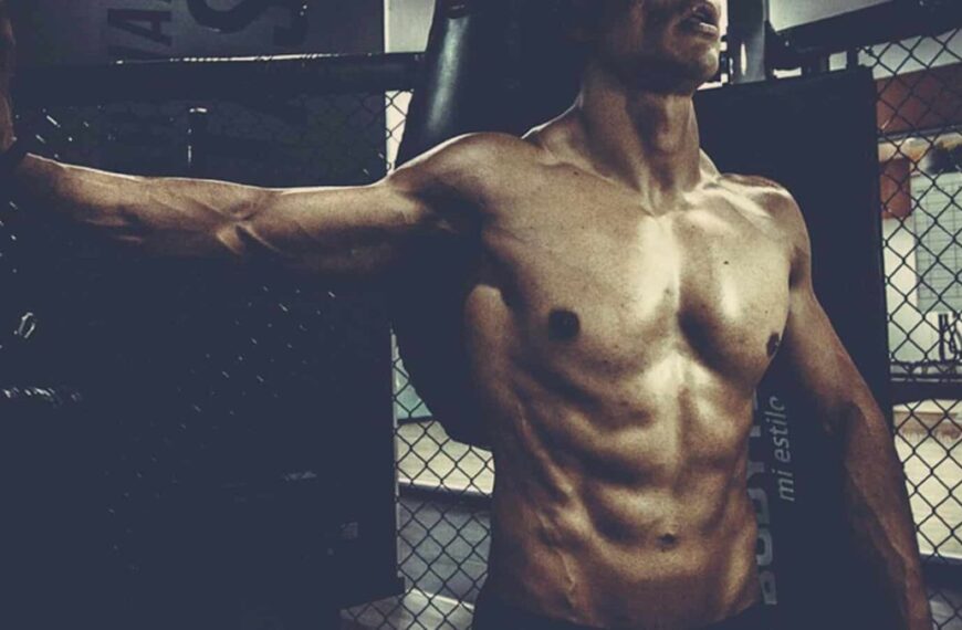 A muscular person stands shirtless in front of a chain-link fence inside a gym, channeling intense focus on sports training, with a punching bag visible in the background.