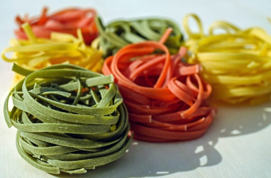 Colorful nests of uncooked pasta in green, red, yellow, and orange, arranged on a light surface—like vibrant passcode keepers guarding the secrets of a well-managed kitchen.