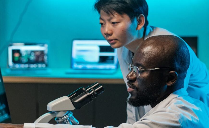 Two scientists in lab coats analyze data on a computer and microscope in a laboratory, exploring emerging approaches to alleviate persistent Lyme disease.