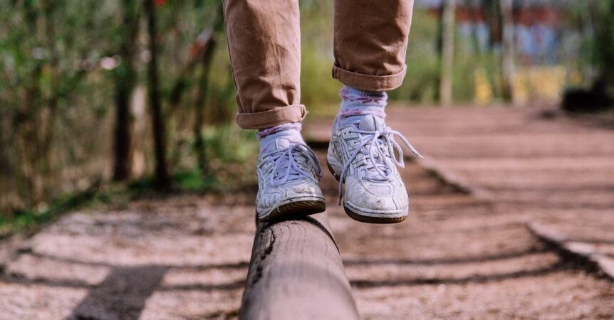Person undergoing vestibular rehabilitation, balancing on a log while wearing white sneakers, patterned socks, and light brown pants on a blurred outdoor path.