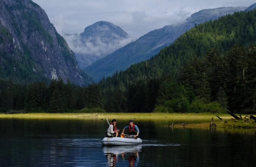 Two people in a small boat paddle across a calm lake, surrounded by lush forests and watched over by forested mountains under a cloudy sky.