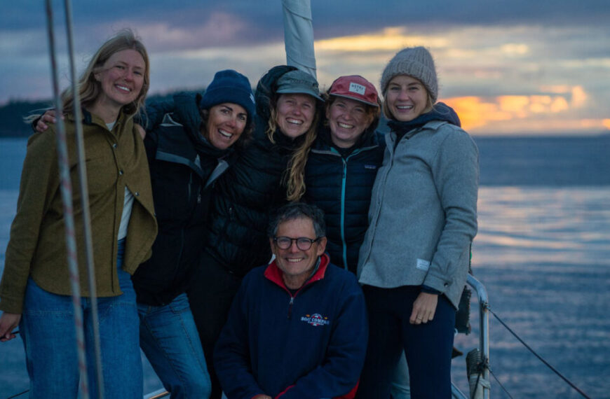 A group of six cheerful helpers smiling on a sailboat at sunset, with a calm sea and colorful sky painting the backdrop.