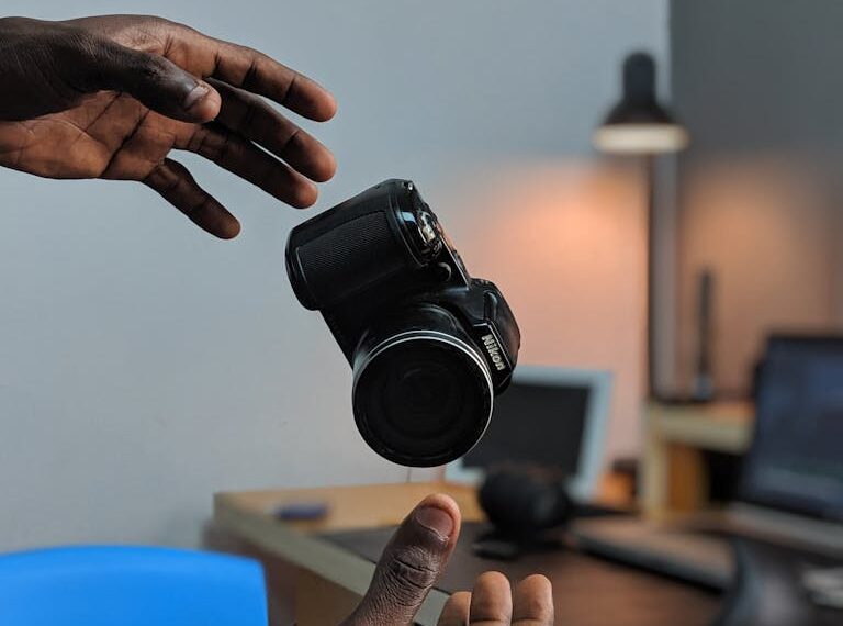 Awkwardly poised between two hands, the camera appears to levitate indoors, with a desk and lamp providing a gentle backdrop.