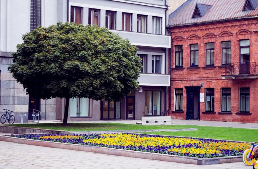 An urban courtyard features a flowerbed of yellow and purple flowers by the entry points, nestled among a large tree and modern brick buildings. Two bicycles are parked nearby, adding charm to this lively scene.
